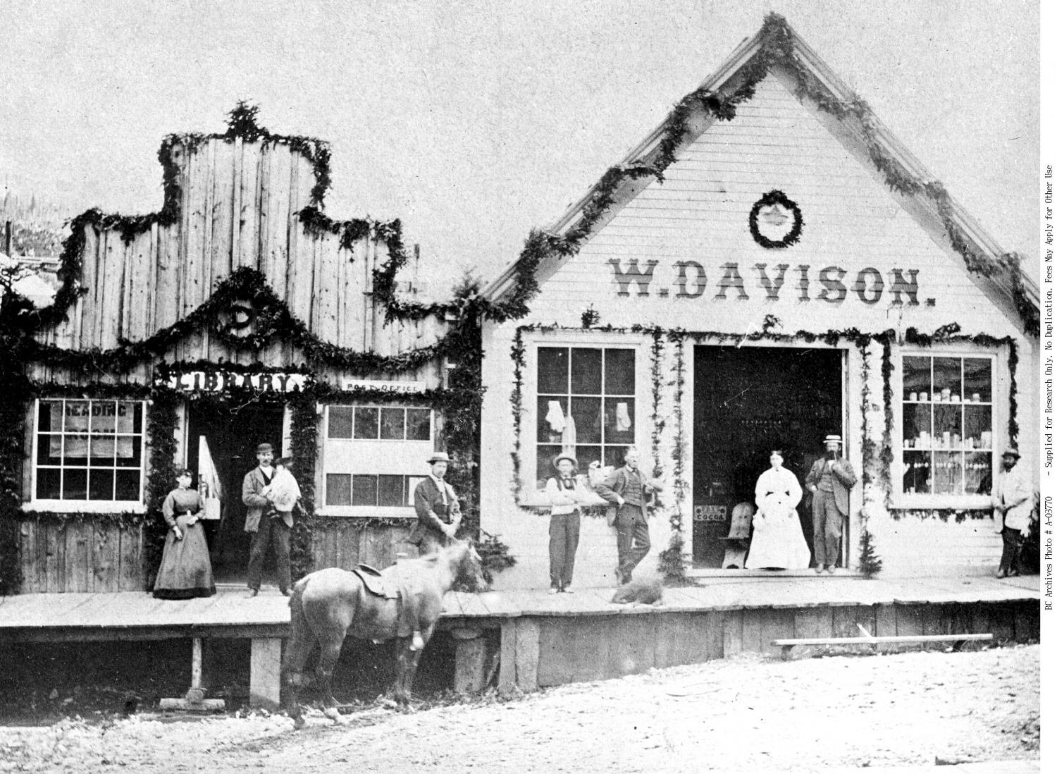 Barkerville Library with Mr. and Mrs. John Bowron in the doorway. Mr. and Mrs. W. Davison in door of grocery shop. Taken in 1871.
