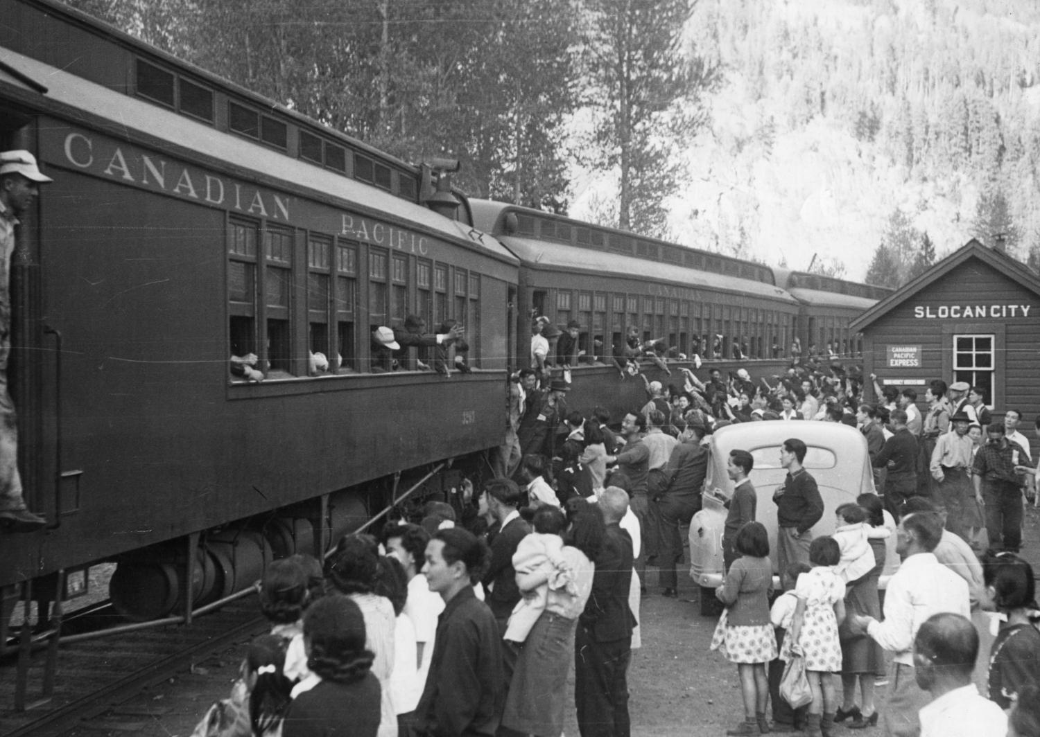 An image of a group of people standing by a train as it departs. A small building in the background has a sign which reads: "Slocan City".