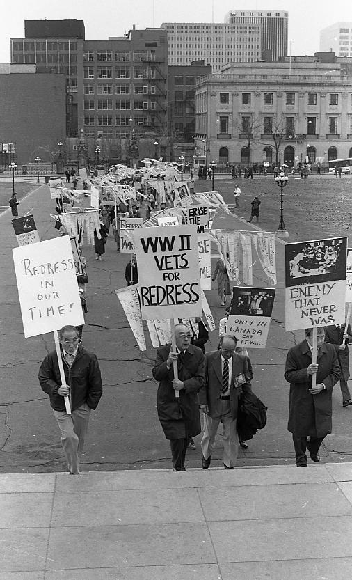 This image shows the Japanese Canadian Redress Rally at Parliament Hill. The placards read: "WWII VETS FOR REDRESS" held by Roger and Mary Obata, "ENEMY THAT NEVER WAS" held by Bill Kobayashi, "LIFE IN EXILE" held by Charles Kadota, "HOME '42"