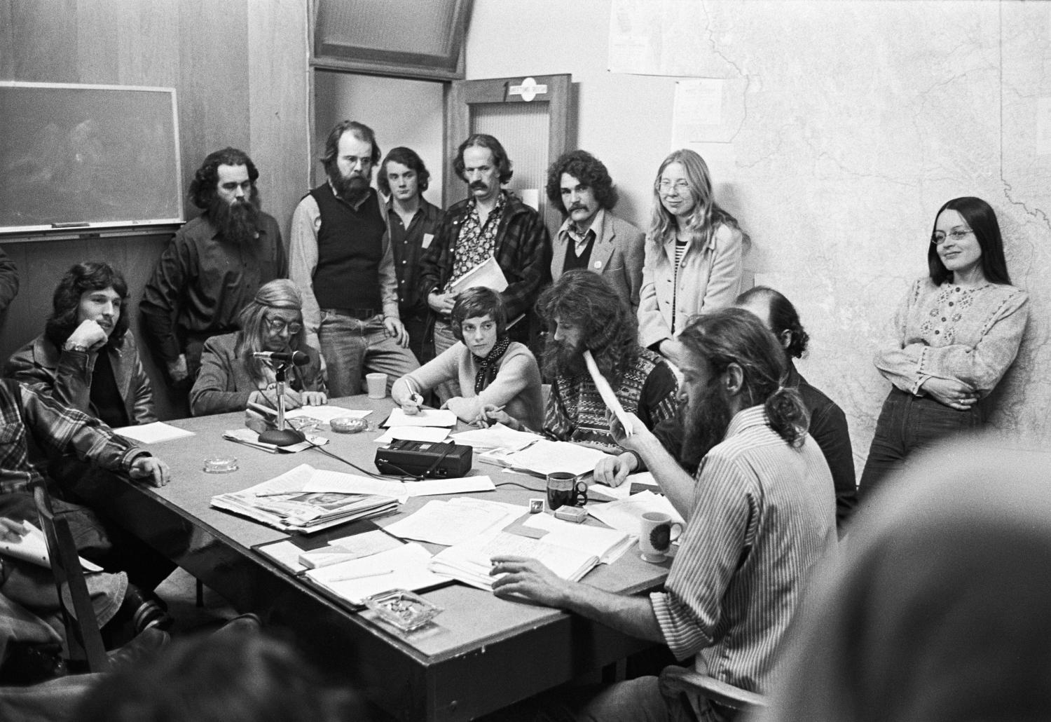 The first public Greenpeace office at 2007 West 4th Avenue in Vancouver, B.C., Canada. Bob Hunter sits at the head of the table, Bree Drummond leans against the wall, Rod Marining, with long red hair and beard sits next to Hunter’s right, Alan Clapp stands in the doorway with a file folder, and Henry Payne sits at the far end of the table with a headband.