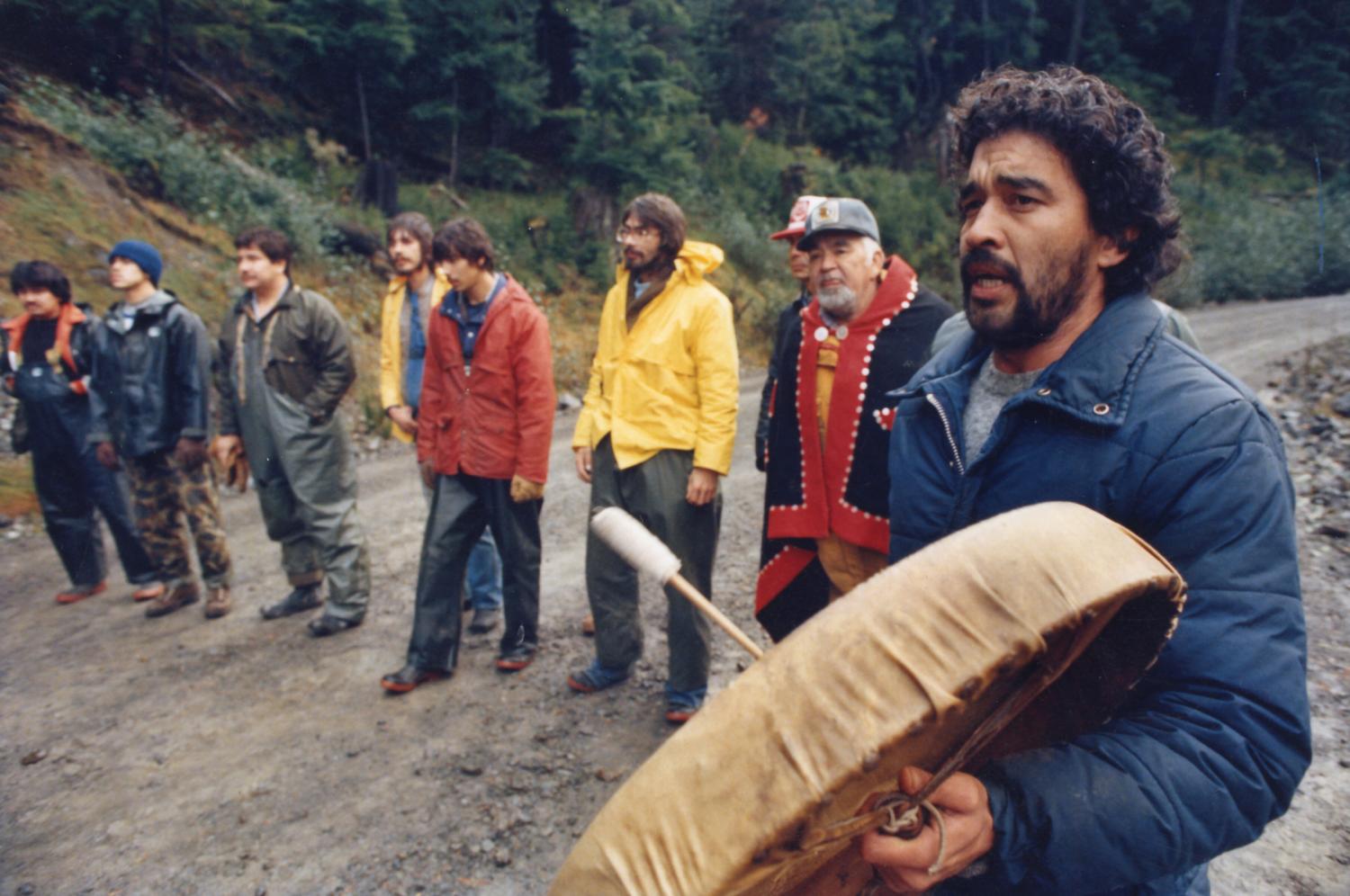 A row of Haida activists and allies block a logging road on Lyell Island. Haida spokesperson Gary Guujaw beats a drum.