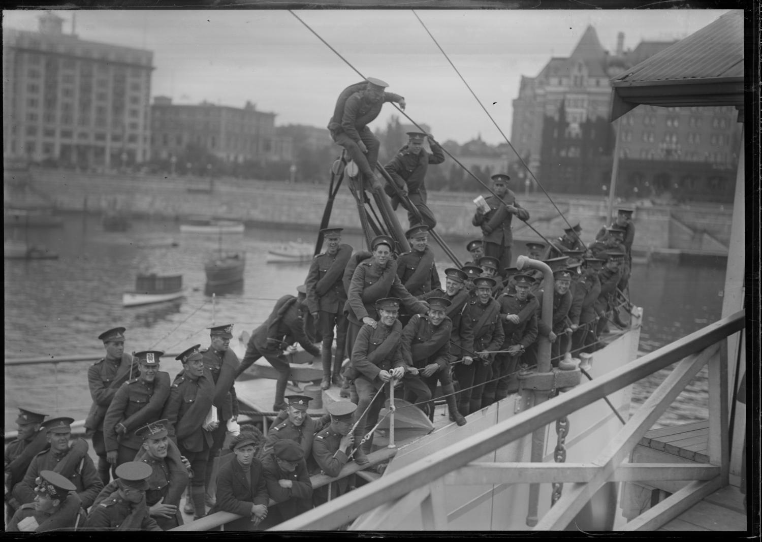 Photograph depicts a draft of soldiers from the Royal Canadian Garrison Artillery. These soldiers are aboard the SS Princess Victoria, a CPR steamship that would transport the soldiers from Victoria to the train station in Vancouver. In this particular shot, some soldiers wear decorated hats and hold care packages. Part of the Empress Hotel can be seen in the background. 
