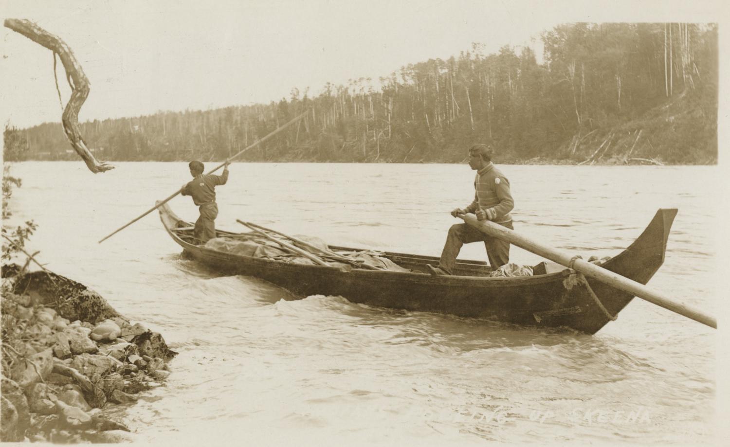 The item is a black and white photograph of the Skeena River taken around 1910 to 1911 by an unknown photographer. The view is from the bank and shows two Indigenous men standing in a canoe. The men are identified as Tsimshian.